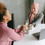 Two women sharing a supportive conversation over coffee indoors. One woman wears a head wrap.