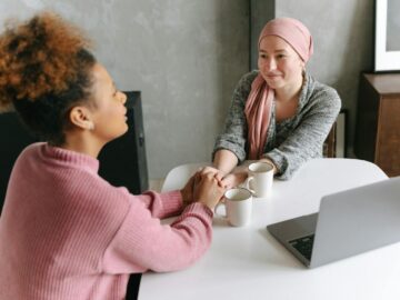 Two women sharing a supportive conversation over coffee indoors. One woman wears a head wrap.