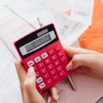 A person calculating finances on a red calculator surrounded by documents.