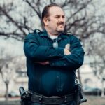 Male police officer with sunglasses standing confidently outdoors in uniform.