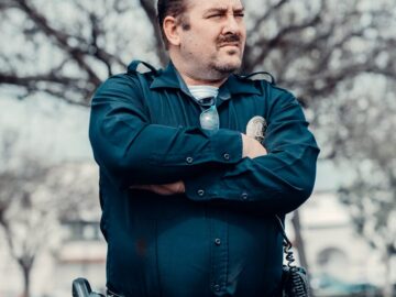 Male police officer with sunglasses standing confidently outdoors in uniform.