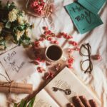 Top view of crop anonymous person with opened book and key placed on white textile with flowers and cup of drink near postcards with inscriptions