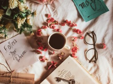 Top view of crop anonymous person with opened book and key placed on white textile with flowers and cup of drink near postcards with inscriptions