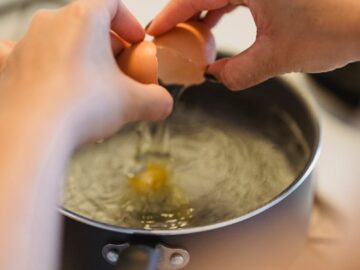 A person cracks an egg into a pot of boiling water, preparing a poached egg.