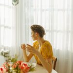 Woman sitting at a sunlit table enjoying a cup of tea, surrounded by a peaceful indoor atmosphere.