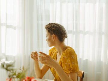 Woman sitting at a sunlit table enjoying a cup of tea, surrounded by a peaceful indoor atmosphere.