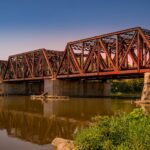 A rusted railway bridge with intricate design reflected in a calm river under clear skies.