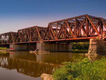 A rusted railway bridge with intricate design reflected in a calm river under clear skies.