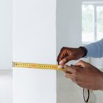 Detailed shot of hands using a tape measure against a white wall indoors.