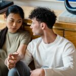 A couple sitting together indoors, holding hands with thoughtful expressions.