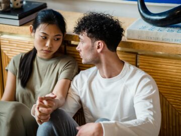 A couple sitting together indoors, holding hands with thoughtful expressions.