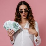 Young woman in sunglasses holding euro banknotes and pointing, pink background