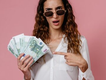 Young woman in sunglasses holding euro banknotes and pointing, pink background