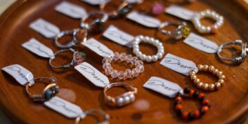 A collection of assorted handmade jewelry rings displayed on a wooden plate with labeled tags.