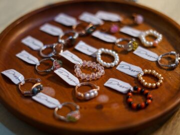 A collection of assorted handmade jewelry rings displayed on a wooden plate with labeled tags.