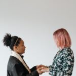 Two women standing indoors holding hands in a prayerful and supportive moment.