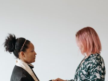 Two women standing indoors holding hands in a prayerful and supportive moment.