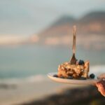 A scenic view of a blueberry pie slice with a fork, held by a hand at a beach.
