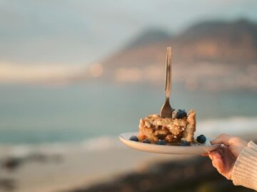 A scenic view of a blueberry pie slice with a fork, held by a hand at a beach.