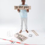 A man stands barefoot in a studio holding a protest sign reading "Do your part" among scattered placards.