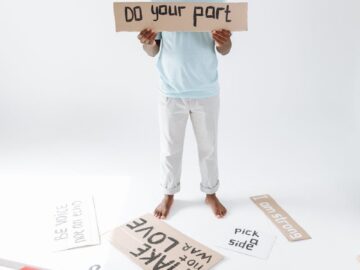 A man stands barefoot in a studio holding a protest sign reading "Do your part" among scattered placards.