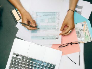 Hands writing on tax documents with laptop, glasses, and currency on desk.