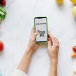 A person using a smartphone for grocery delivery surrounded by fresh fruits and vegetables.