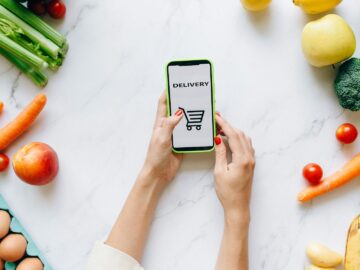 A person using a smartphone for grocery delivery surrounded by fresh fruits and vegetables.