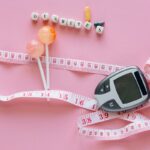 Close-up of a diabetes glucometer, tape measure, and pills on pink surface.