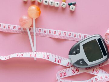 Close-up of a diabetes glucometer, tape measure, and pills on pink surface.