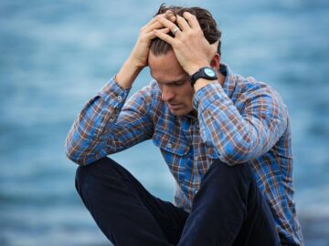 A man in a plaid shirt sits by the water looking distressed, symbolizing stress.