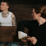 A tattooed man in a white tank top sits opposite a priest holding a holy book, symbolizing a contrast of lifestyles and beliefs.