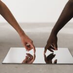 Two hands of different ethnicities touching and reflecting on a minimalist mirror.