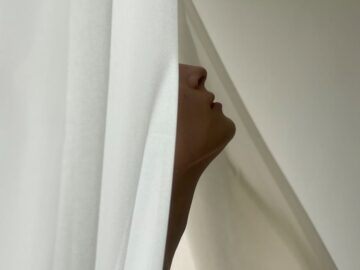 Intriguing profile of a woman partially hidden by a white curtain, captured in soft lighting.